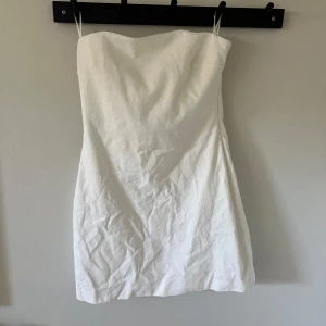 Vit axelbandslös kort klänning - Säljer en stilren vit axelbandslös kort klänning i bomull. Klänningen har en enkel och rak passform som sitter snyggt på kroppen. Perfekt för sommarens varma dagar eller när du vill ha en clean look. Lätt att matcha med accessoarer och sneakers. Har en liten missfärgning längst ner, har ej försökt få bort.