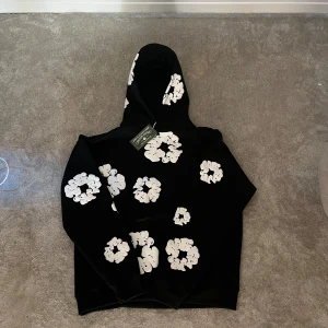 Svart hoodie från DT - Svart hoodie från DT med stora vita blommor tryckta över hela tröjan. Tröjan har huva och en avslappnad passform, perfekt för dig som gillar streetwear med unik design. Materialet känns mjukt och skönt mot huden.