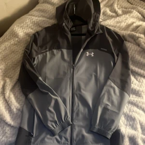 Grå vindjacka från Under Armour köpte den för några dgr sen o jag tycker inte om designen så säljer den knappast använd  - Snygg grå vindjacka från Under Armour med huva och dragkedja. Jackan har en stilren design med logga på bröstet och stor text på ryggen. Tillverkad i polyester och perfekt för blåsiga dagar. Elastiska muddar vid ärmslut och praktiska fickor. 