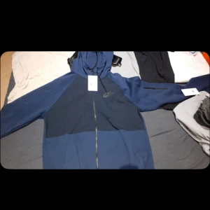 Blå Nike tech tröja stl M  - Snygg tvåfärgad blå vindjacka från Nike med hel dragkedja och huva. Jackan har svarta detaljer och en diskret Nike-logga på bröstet. Perfekt för dig som gillar sportig stil och vill ha något bekvämt och stilrent. Säljer för den inte passar 