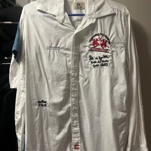 Vit skjorta från La Martina - Vit långärmad skjorta från La Martina med broderade detaljer och logga på bröstet samt stor text på ryggen. Skjortan har klassisk krage, knäppning framtill och är tillverkad i bomull. Snygg och sportig stil med unika broderier och detaljer.