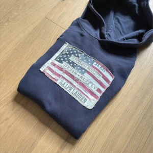 Mörkblå hoodie Ralph Lauren flag patch - Snygg mörkblå hoodie från Ralph Lauren Denim & Supply med stor amerikansk flag patch framtill. Tröjan har huva och är tillverkad i mjuk bomull, perfekt för chill dagar. Ikonisk streetstil med vintage-vibe och coola detaljer.