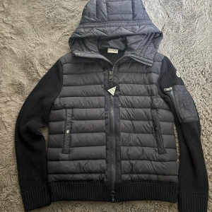 Navy Moncler cardigan - Snygg navy pufferjacka från Moncler med stickade ärmar och vadderad kropp. Jackan har huva, dragkedja framtill och praktiska fickor. Ärmarna är ribbstickade och har en Moncler-logga på ena armen. Perfekt för kyliga dagar och riktigt stilren look. Helt Ny säljer för den inte passade mig.