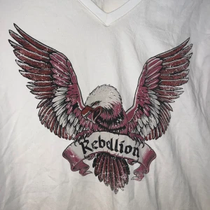 Vit t-shirt med örntryck från Culture - Vit mc liknande t-shirt från Culture med v-ringning och korta ärmar. Framsidan har ett stort tryck av en örn med utbredda vingar och texten 'Rebellion' i gotisk stil. Materialet är mjuk bomull och passformen är normal. Perfekt för dig som gillar edgy och coola prints. Aldrig använd. Kan va en oversized S eller perfekt L