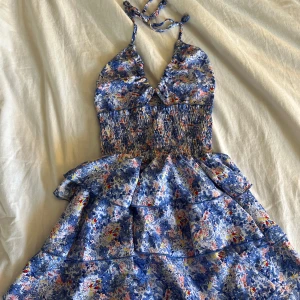 Blommig blå kortklänning med volanger shein - Supersöt kortklänning med blått blommönster, smockad midja och volanger i kjolen. Klänningen har halterneck med knyt i nacken och v-ringad urringning. Perfekt för sommarens alla tillfällen och ger en riktigt fräsch vibe.