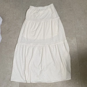 Vit långkjol med broderade detaljer Kappahl - Superfin vit långkjol från Kappahl med broderade hål och volangiga partier. Kjolen har resår i midjan och är gjord i ett lätt och luftigt bomullstyg, perfekt för varma dagar. Snygg och enkel att matcha med en crop top eller t-shirt.