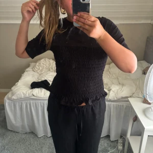 Svart smockad topp med volangkant - Säljer en svart topp med smockad midja och volangkant nertill. Toppen har korta ärmar och en figurnära passform som framhäver midjan. Perfekt att styla med både jeans och kjol för en trendig look.