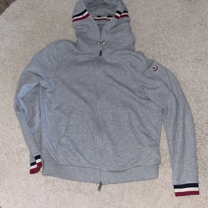 Grå Moncler hoodie med huva - Snygg grå hoodie från Moncler med hel dragkedja, huva och Moncler-logga på ärmen. Ribbade muddar med röd, vit och marinblå rand. Perfekt för chill dagar och streetstyle. Tillverkad i mjuk bomull för maximal komfort. Passform är M - L