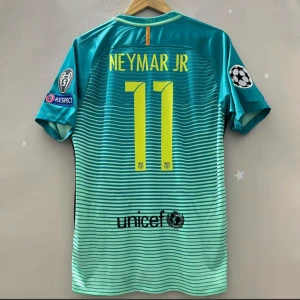 FC Barcelona Neymar Jr 11 matchtröja - Snygg FC Barcelona fotbollströja med Neymar Jr och nummer 11 på ryggen. Tröjan är turkosblå med tunna horisontella ränder, gula detaljer och officiella Champions League-patchar på ärmarna. Tillverkad i lätt och ventilerande material, perfekt för fotbollsälskare.