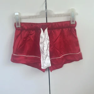Pyjamasshorts röda i satin från Gina Tricot - Snygga röda satinskjorts från Gina Tricot med vita detaljer och dekorativ vit snörning framtill. De har elastisk midja och vita passpoaler längs benen för en sportig vibe. Perfekta för sommarens chill eller som pyjamasshorts.