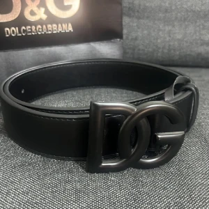 Svart skärp  - Stilrent svart bälte med bred rem i skinn och en stor, mattsvart DG-logga som spänne. Kommer med originalpåse och ask, perfekt för dig som vill ha en lyxig accessoar med tydlig designerstil.