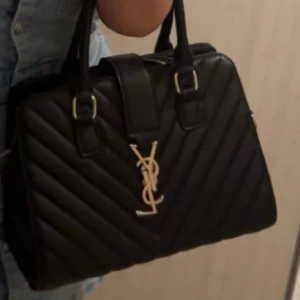 Svart quiltad handväska från Saint Laurent - Säljer en svart quiltad handväska från Saint Laurent med guldiga detaljer och klassiskt YSL-emblem framtill. Väskan är tillverkad i skinn och har två rejäla handtag samt dragkedja upptill. Perfekt för dig som vill ha en lyxig och stilren accessoar.