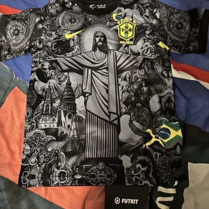 Brasil Nike fotbollströja svart/gul - Nytt skick. Snygg Brasilien fotbollströja från Nike med unikt svart och grått grafiskt mönster, stora tryck av Kristusstatyn och brasilianska symboler. Gula detaljer och CBF-emblem på bröstet. Tillverkad i lätt och ventilerande material, perfekt för match eller träning. Man får shorts och t-shirten. Priset kan diskuteras!
