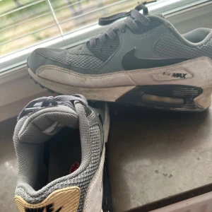 Nike Air Max grå sneakers - Säljer ett par klassiska Nike Air Max sneakers i grått med svarta detaljer och vit sula. Ovandelen är i mesh och syntet, med tydlig Air Max-dämpning i sulan. Skorna har snörning och rund tå, samt beige detalj vid hälen med Nike-logga.