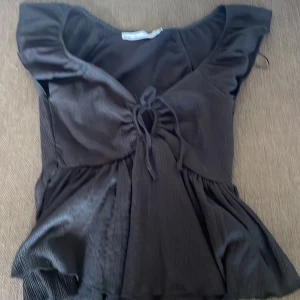 Svart blus med volang och knyt - Supersöt svart blus med djup v-ringning, volangärmar och knyt framtill. Modellen är lätt utställd och har en flowy känsla. Perfekt till jeans eller kjol för en trendig look. Gjord i ett mjukt, lätt tyg som känns skönt mot huden.