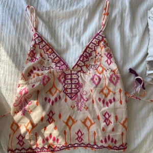 Mönstrad linnetopp med snörning - Superfin linnetopp med smala axelband och färgglatt mönster i rosa, orange och lila. Toppen har v-ringning, broderade detaljer och snörning framtill samt knytband i midjan med tofsar. Perfekt för varma dagar och festivaler.