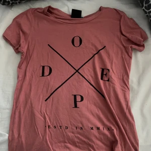 Rosa t-shirt med svart tryck från Dope - Snygg rosa t-shirt från Dope med svart grafiskt tryck på framsidan. T-shirten har rund halsringning och korta ärmar. 