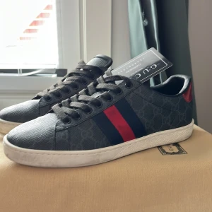 Gucci ace skor - Säljer ett par gråa sneakers från Gucci med klassiska röda och marinblå ränder på sidan. Skorna har svart snörning, diskret mönster i tyget och vit platt sula. Röd detalj vid hälen. Perfekta för en stilren vardagslook.