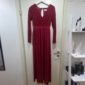 Röd långklänning med spets och v-ringning från Chiara Forthi - Elegant röd långklänning med v-ringning och spetsdetaljer upptill. Klänningen har långa ärmar i spets och en markerad midja. Baksidan har dekorativa knappar och en öppen detalj. Perfekt för bal eller fest.