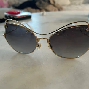 solglasögon med guldiga bågar - Unika solglasögon med guldiga metallbågar och svarta till gråtonade glas. Glasen har en vågig, nästan hjärtformad design och tunna vita skalmar. Perfekt för dig som vill sticka ut med en cool accessoar. Med Rostigt