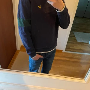 Mörkblå sweatshirt från Lyle & Scott - Säljer en mörkblå sweatshirt från Lyle & Scott med gröna detaljer på ärmarna och den klassiska gula loggan på bröstet. Tröjan har rund hals och långa ärmar. Storlek S. Använd ett flertal gånger och det är ett pyttelitet hål vid höger om halsen