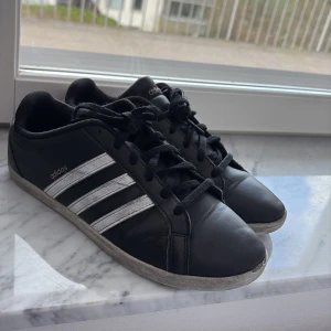 Svarta Adidas sneakers med vita ränder - Klassiska svarta Adidas sneakers med tre vita ränder på sidorna och svart snörning. Skorna har en låg profil och platt sula, perfekt för en stilren vardagslook. Adidas-logga i vitt på sidan och tungan.