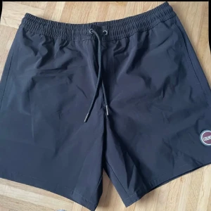 Svarta badshorts från italienska Colmar 48 - Snygga svarta badshorts från Colmar med elastisk midja och snörning. Shortsen har en bakficka med lock och Colmar-logga både fram och bak. Perfekta för stranden eller poolen.
