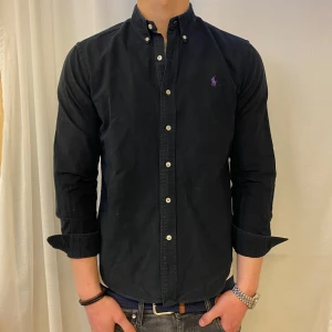 Svart skjorta från Polo Ralph Lauren - Säljer en svart långärmad skjorta från Polo Ralph Lauren i slim fit-modell. Skjortan har klassisk button down-krage, vita knappar och lila broderad logga på bröstet. Tillverkad i stretchig oxford-bomull för extra komfort.