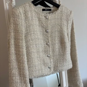 Vit kavajjacka från Gina Tricot - Säljer en elegant vit kavajjacka från Gina Tricot i storlek 32. Jackan har ett glittrigt tyg, rund halsringning och dekorativa silverfärgade knappar framtill. Perfekt för att lyfta vilken outfit som helst.