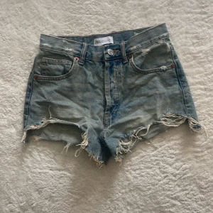 Jeansshorts från Zara - Säljer ett par ljusblå jeansshorts från ZARA med fransiga kanter och slitna detaljer. Klassisk femficksmodell med knapp och dragkedja framtill. Perfekta för varma dagar!