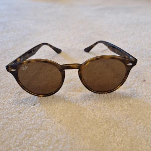 Bruna Ray-Ban 2180  - Snygga solglasögon från Ray-Ban med rund form och brunmelerat mönster på bågarna. Original pris 1 800. Glasen är mörka och bågarna har en klassisk look med små silvriga detaljer vid fronten. Perfekt accessoar för soliga dagar.