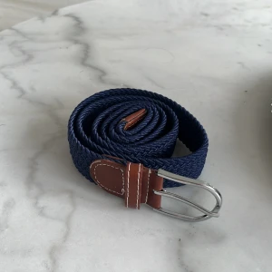 Flätat bälte - Snyggt mörkblått flätat bälte med bruna skinn-detaljer och silverfärgat spänne. Perfekt accessoar för att lyfta din outfit. Bältet har en klassisk design och passar till många olika stilar.