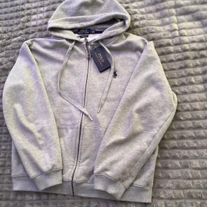 Zip hoodie Polo Ralph Lauren - Säljer en klassisk grå hoodie från Polo Ralph Lauren med dragkedja och huva. Tröjan har ribbade muddar, fickor framtill och den ikoniska broderade loggan på bröstet. Perfekt för en avslappnad stil.