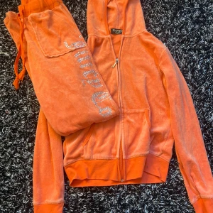 Orange mjukisbyxor från Juicy Couture - Säljer ett par orange mjukisbyxor från Juicy Couture i mjuk velour med glittrig Juicy-text och krona på baksidan. Byxorna har resår i midjan och fickor med knapp bak. Perfekt för en chill och trendig look.