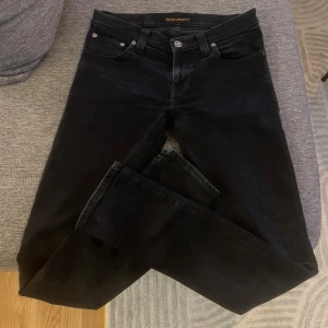  Nudie Jeans - Säljer ett par svarta jeans från Nudie Jeans med klassisk femficksdesign och diskret broderad bakficka. Jeansen har silverfärgade knappar och raka ben. Perfekta för dig som gillar stilrena och tidlösa plagg.