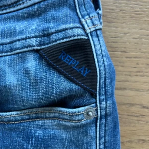 Blå Replay jeans Anbass - Säljer ett par klassiska blå jeans från Replay med raka ben och snygga slitningar. Jeansen har fem fickor, silverfärgade nitar och svart Replay-läderpatch bak i midjan. Perfekta för en avslappnad stil.