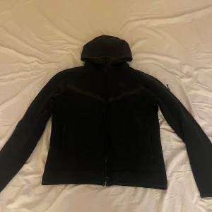 Svart hoodiejacka från Nike - Säljer en svart hoodiejacka från Nike med dragkedja framtill. Jackan har huva och är långärmad, perfekt för en sportig och avslappnad stil. Passar dig som gillar enkel och stilren design.