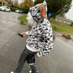 Vit och svart camo hoodie från BAPE - Säljer en hoodie från BAPE i vit och svart camo.. Dragkedja hela vägen upp och broderade detaljer i färg. Perfekt för dig som vill sticka ut med streetwear-stil. Lyser även i mörkret