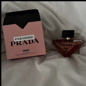Paradoxe Intense Eau de Parfum från Prada - Paradoxe Intense från Prada är en lyxig parfym i en unik trekantig glasflaska med svart lock. Förpackningen är rosa med svart och vit text. Doften är intensiv och elegant, perfekt för dig som vill sticka ut med en modern och exklusiv parfym.