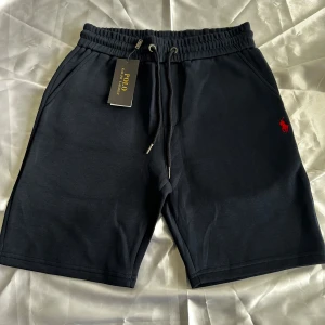 Svarta shorts från Polo Ralph Lauren - Svarta shorts från Polo Ralph Lauren med elastisk midja och snörning. Klassisk röd logga broderad på vänster ben och fickor på sidorna samt en bakficka. Perfekta för en avslappnad stil.