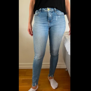 Ljusblå skinny jeans med slitningar - Säljer ett par ljusblå skinny jeans med hög midja och slitna detaljer vid knä och benslut. Jeansen har klassisk femficksdesign, dragkedja och knapp framtill. Perfekta för en avslappnad och trendig look.