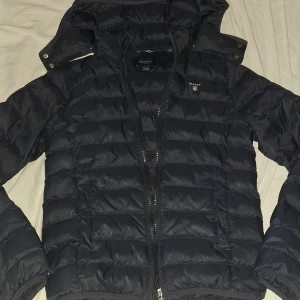 Svart pufferjacka från Gant med huva - Svart pufferjacka från Gant med avtagbar huva och dragkedja framtill. Jackan har quiltad design och Gant-logga på bröstet. Perfekt för kyligare dagar och har en sportig look. Nått litet småhåll här och där men annars bra skick! Pris kan diskuteras.