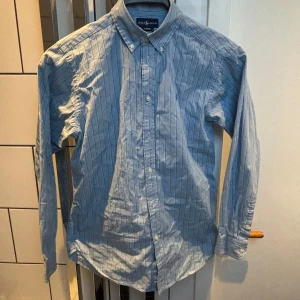 Ralph Lauren skjorta - Säljer en klassisk blå skjorta från Ralph Lauren med vita ränder och broderad logga på bröstet. Skjortan har lång ärm, knappar hela vägen och button-down krage. Perfekt för dig som gillar stilrena plagg med snygga detaljer. Pris kan diskuteras.