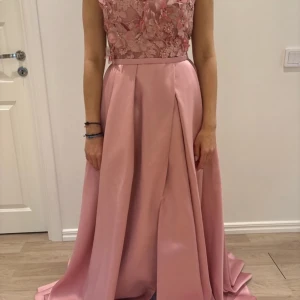 Rosa balklänning med spets och puffärmar - Elegant långklänning i ljusrosa med vacker spets upptill och puffärmar. Kjolen är i satinliknande material med slits framtill och markerad midja. Perfekt för bal eller festliga tillfällen. Är i storlek xs/s ifrån rever design ca 140 cm längd endas använd en gång 