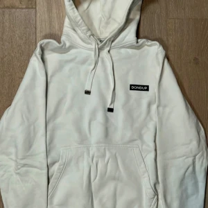 Vit hoodie från Dondup - Säljer en stilren vit hoodie från Dondup med svart logotyp på bröstet. Tröjan har huva med dragsko och en stor magficka framtill. Perfekt för en avslappnad och trendig look.