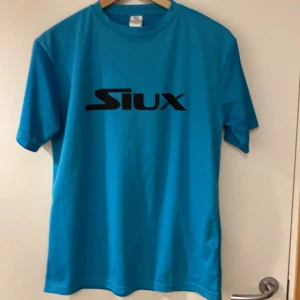 Blå t-shirt från Siux large - Säljer en blå t-shirt från Siux med svart logga tryckt på bröstet. T-shirten har rund halsringning och korta ärmar. Perfekt för dig som gillar sportig stil.