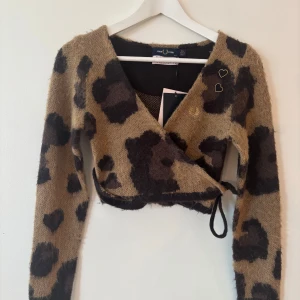 Leopardmönstrad omlottopp från Fred Perry x Amy Winehouse - Säljer en superfin fluffig, croppad omlottopp från Fred Perry x Amy Winehouse med leopardmönster i beige och svart. Tröjan har långa ärmar, broderade hjärtan och knyt i sidan. Helt ny med tags. Nypris ligger på 2200kr. 