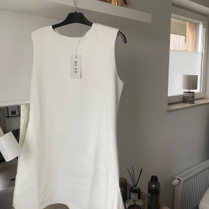 Vit asymmetrisk klänning från NA-KD - Säljer en elegant vitastmetrisl klänning med sidosläp från NA-KD. Klänningen har ärmlös design och en stilren, rak passform med lätt utsvängd nederdel. Perfekt för sommarens tillfällen när du vill känna dig extra fin. Otroligt chic💘