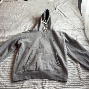 Grå hoodie från Tommy Hilfiger - Säljer en klassisk grå hoodie från Tommy Hilfiger med vita snören och liten logga på bröstet. Tröjan har känguruficka och ribbade muddar vid ärmslut och nederkant. Perfekt för en avslappnad stil.