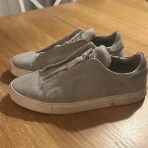 Ljusgrå sneakers med dragkedja från Axel Arigato - Säljer ett par ljusgrå sneakers från Axel Arigato i mocka med dragkedja. Skorna har rund tå och vit platt sula, perfekt för en clean och stilren look. Vid frågor eller funderingar är det bara att höra av sig! Tvättas innan frakt!